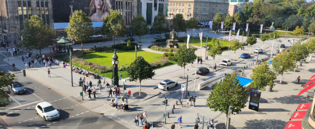 Das Bild zeigt den Corneliusplatz in Düsseldorf. In der Mitte eine gepflegte, formal mit Brunnen gestaltete Grünfläche. Drumherum parken Autos.